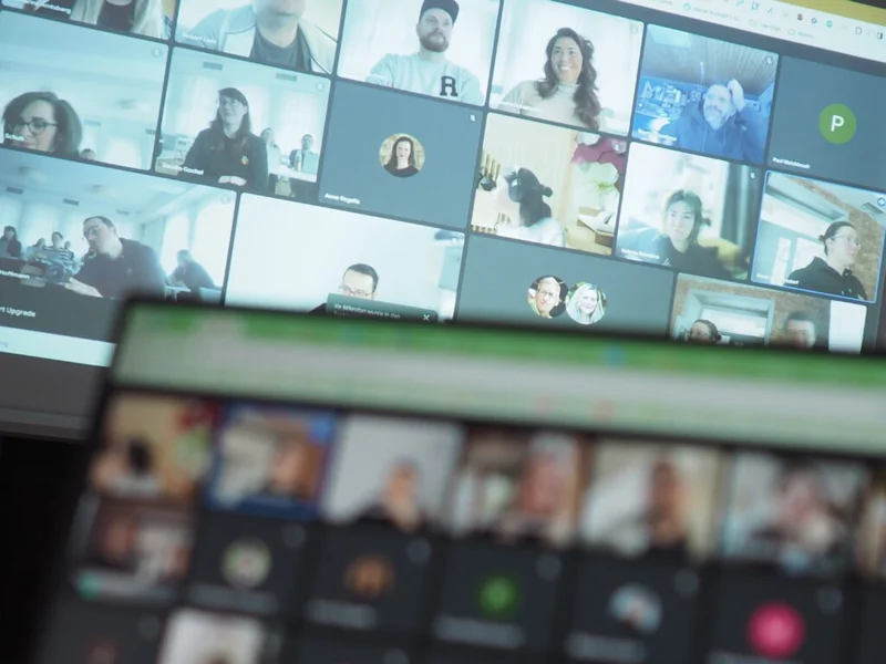 Unscharfes Foto eines Computerbildschirms mit einer Videokonferenz, bei der mehrere Teilnehmer in verschiedenen Umgebungen sichtbar sind. Einige Personen sind einzeln, andere zusammen in Gruppen dargestellt. Der Vordergrund zeigt verschwommene Tasten eines Laptops, was darauf hinweist, dass das Bild von einem Teilnehmer der Videokonferenz aufgenommen wurde.
